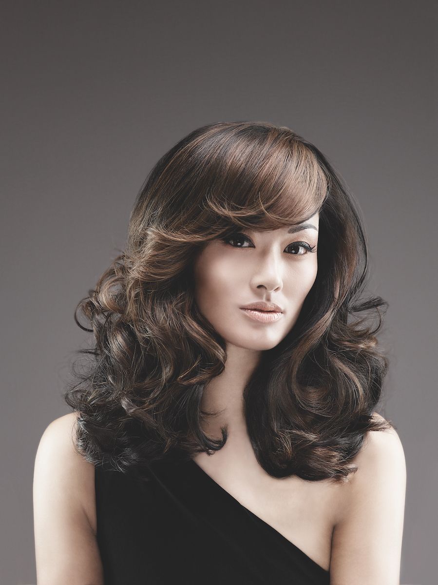 Portrait of a woman with long wavy dark hair in a black one-shoulder top against a gray background
