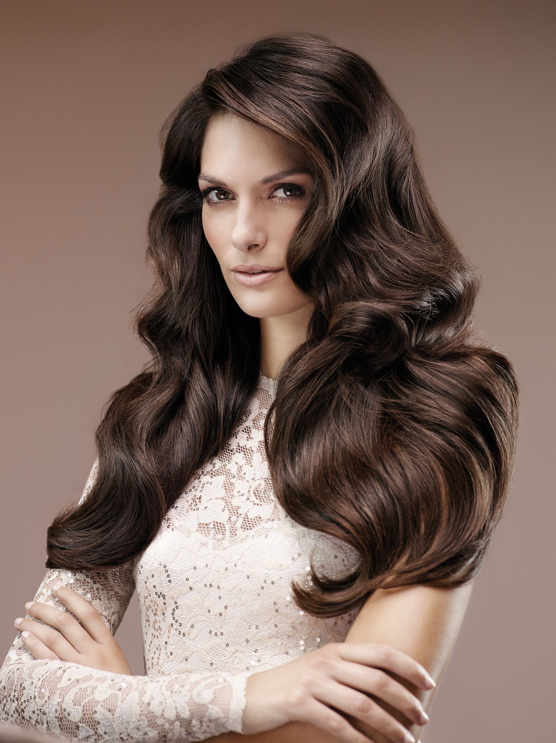 Portrait of a woman with long wavy brown hair in a white lace top, posing against a neutral background