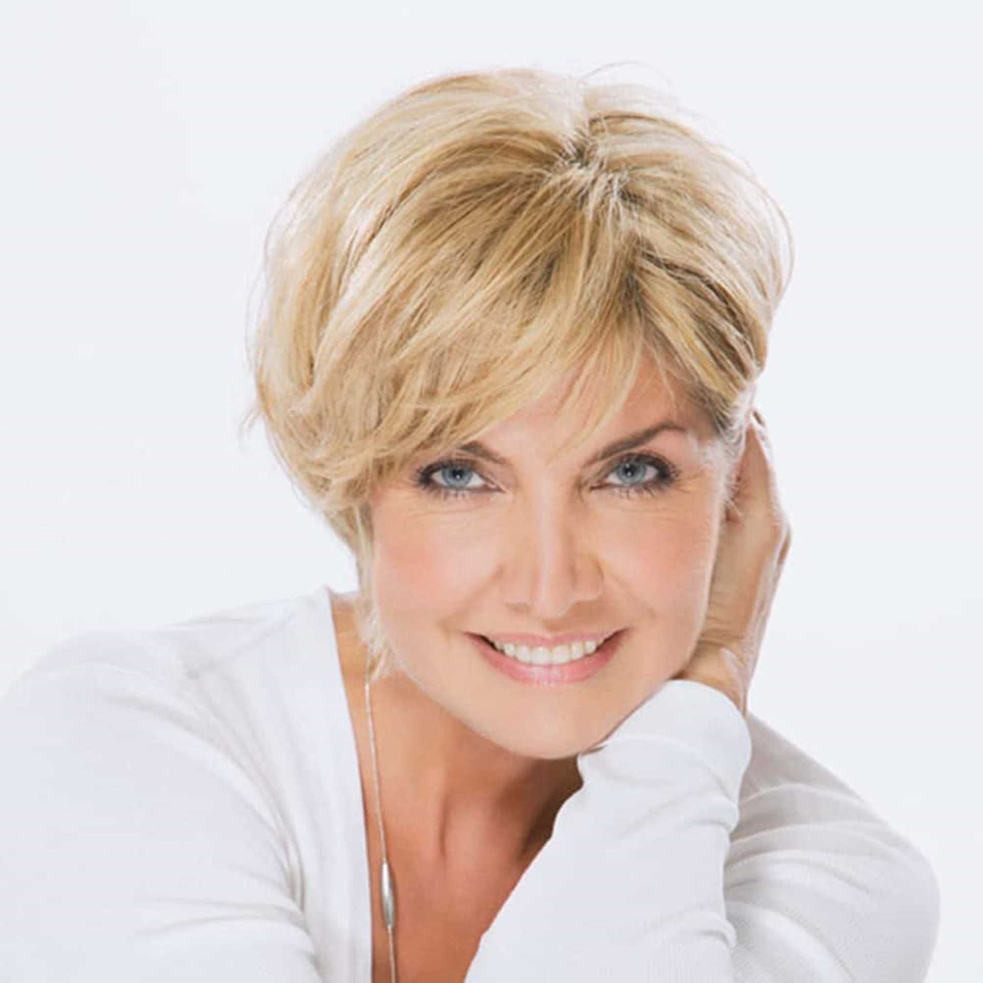 Smiling blonde woman in a white top resting her head on her hand against a white background