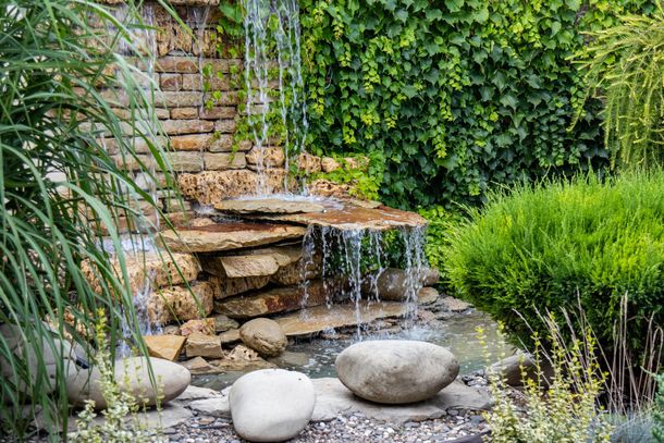 Una piccola cascata da giardino che si riversa su pietre impilate in uno stagno di ciottoli, circondato da una vegetazione lussureggiante.