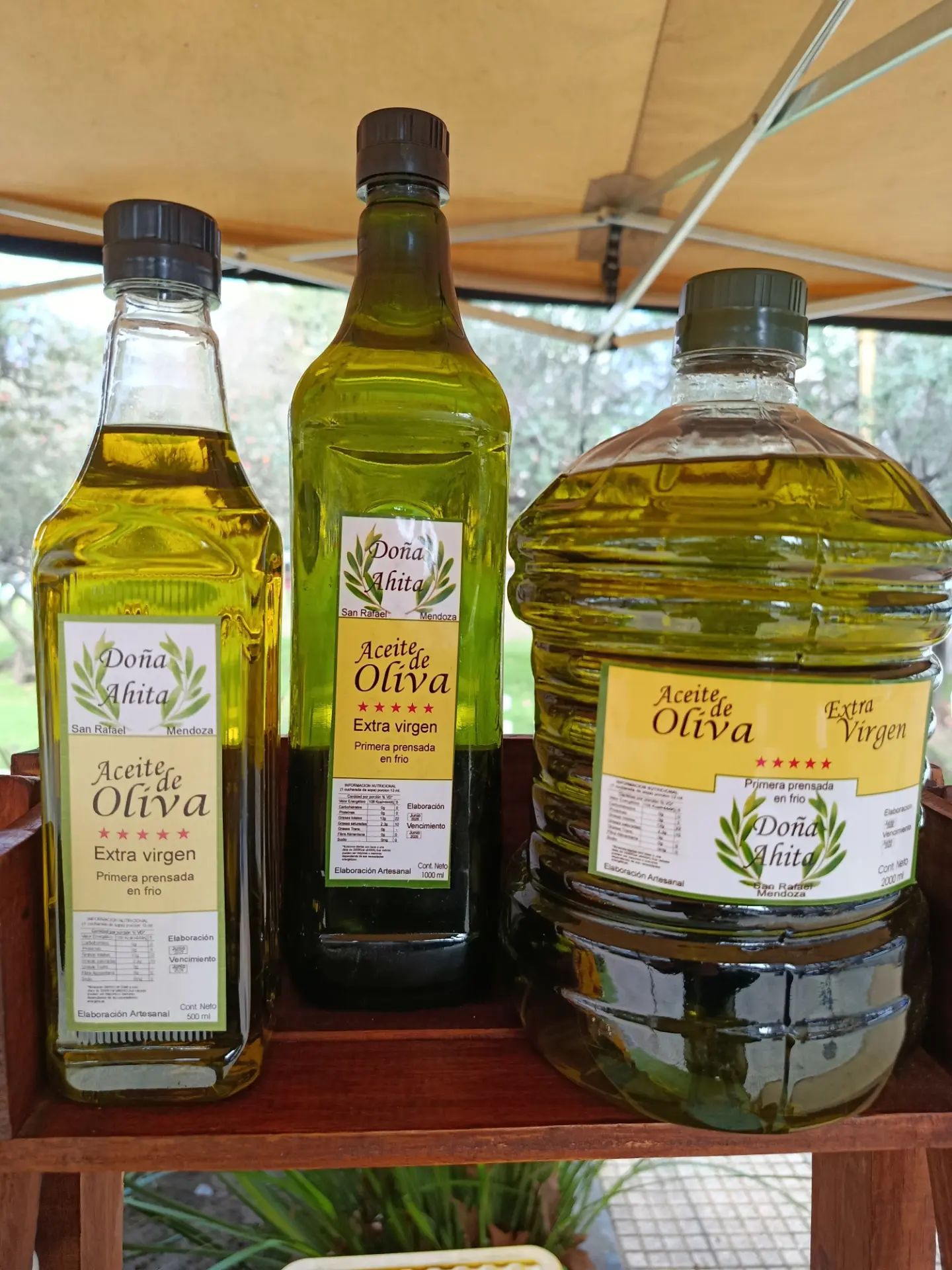 Tres botellas de aceite de oliva sobre una estantería de madera bajo un toldo, con etiquetas verdes y amarillas.