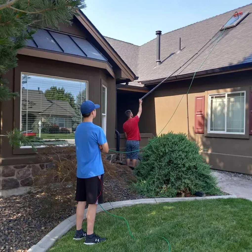 Two men cleaning a house. One uses a long tool on the roof, another watches from the lawn.
