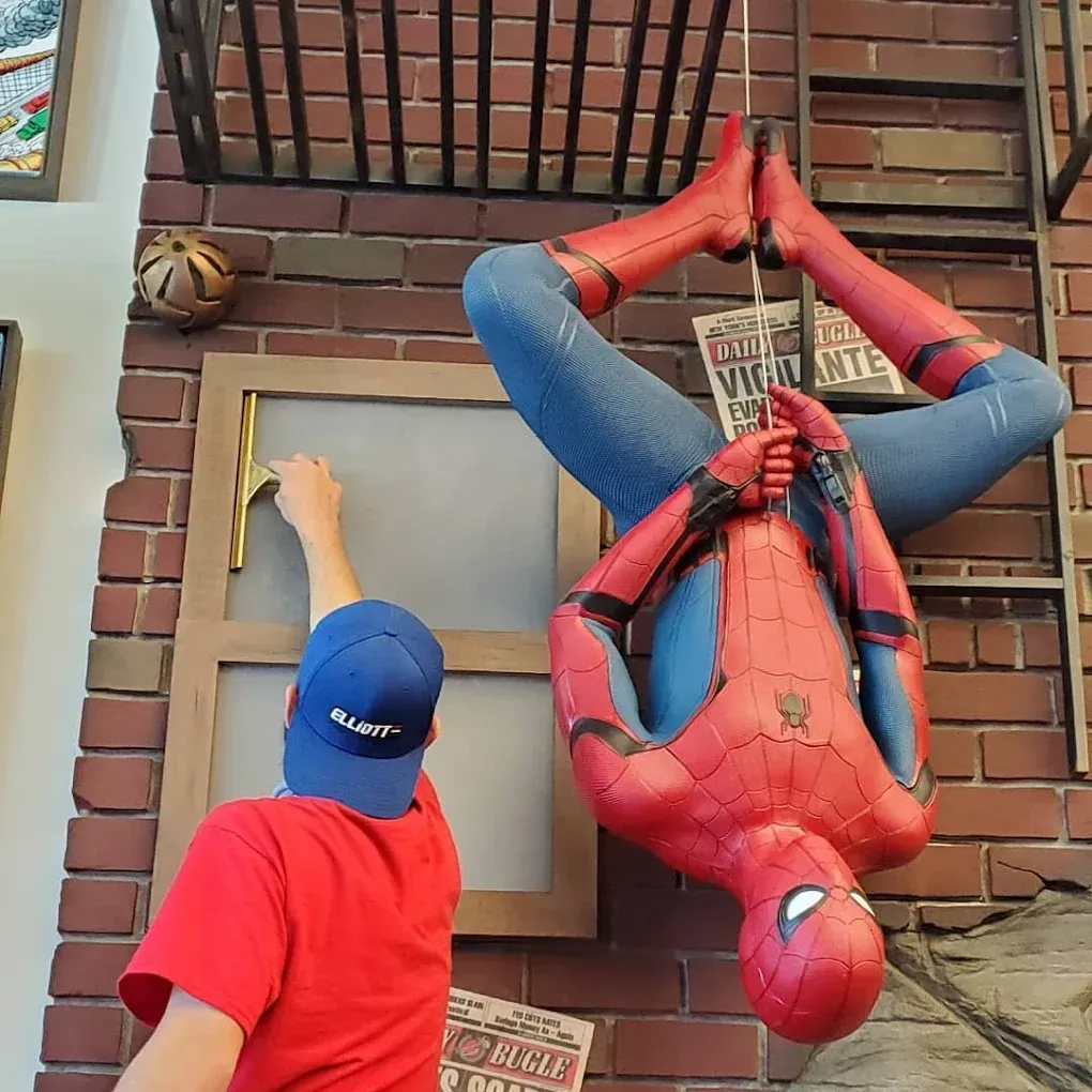 Person in red shirt interacting with Spider-Man statue hanging upside down on a brick wall, next to a window.