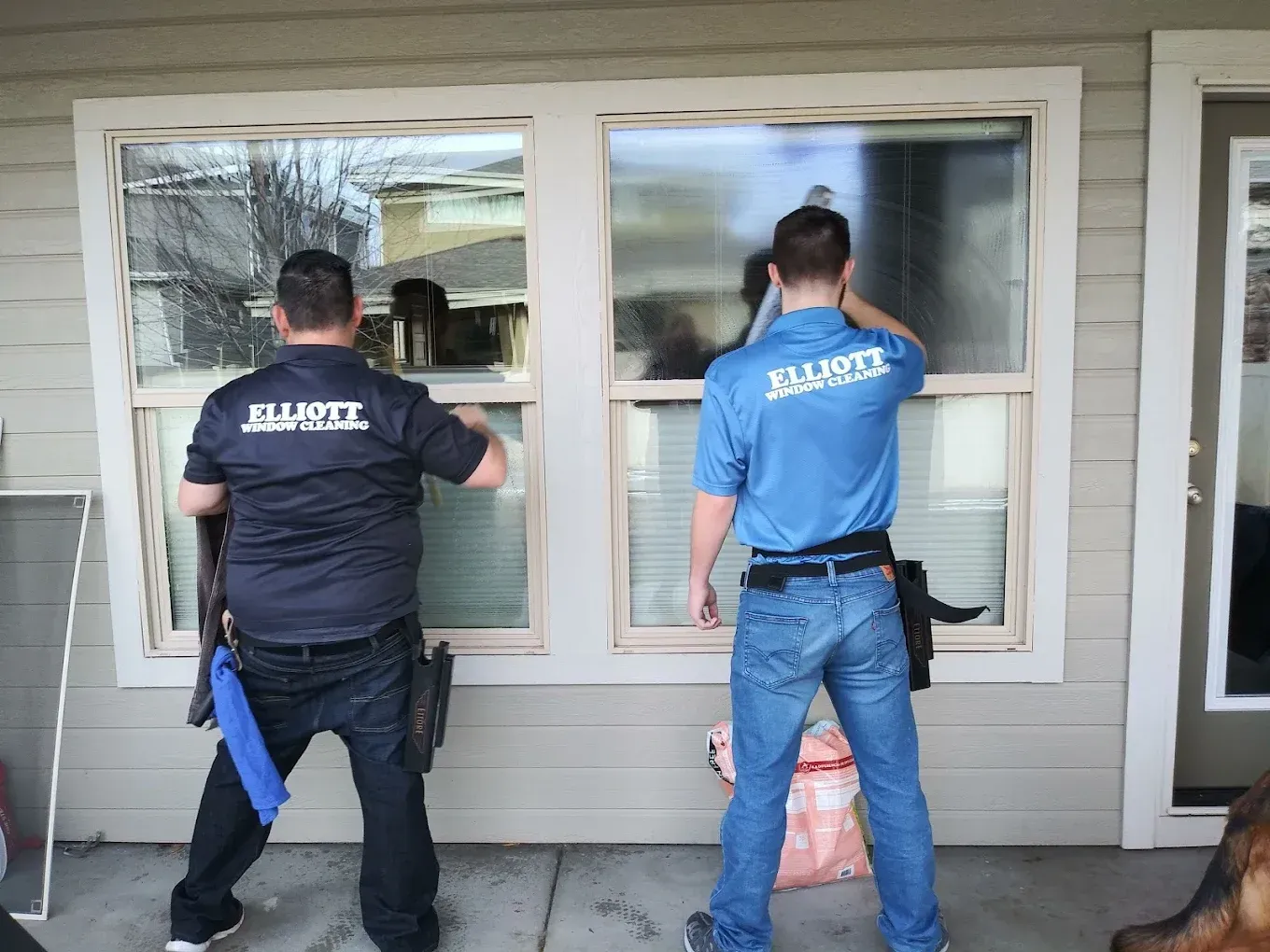 Two men in uniform cleaning windows of a house. One in black, one in blue. Exterior shot.