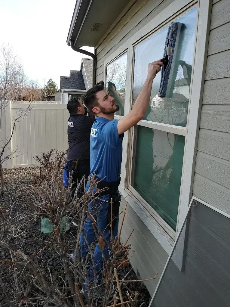 Two men washing a window on a house. One uses a squeegee. Outdoors, sunny day.