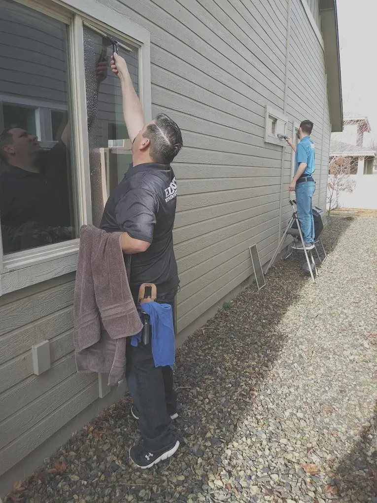 Two people cleaning a window on the side of a house. One person is inside, and the other is outside on a ladder.