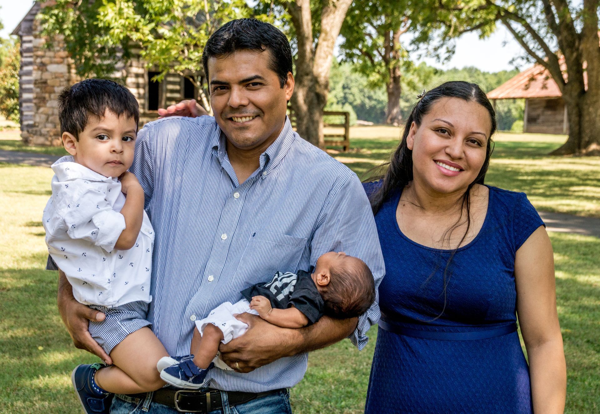 Family of four smiling outside: man holding two young children, woman beside them.