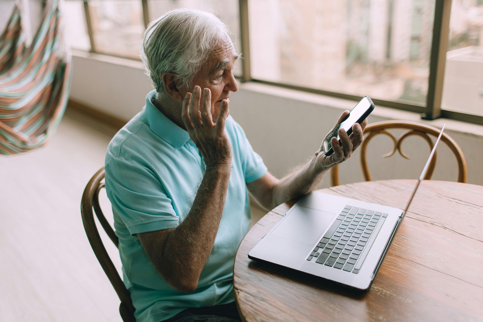 Elderly man in blue shirt, holding phone, smiling, looking at laptop on table near a window.