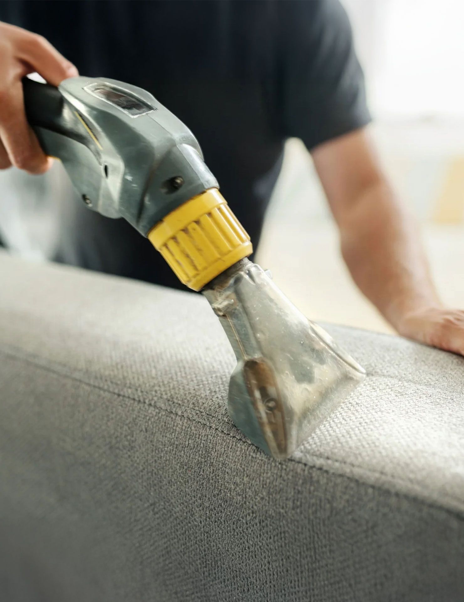 A person uses a spray upholstery cleaner on a gray sofa.