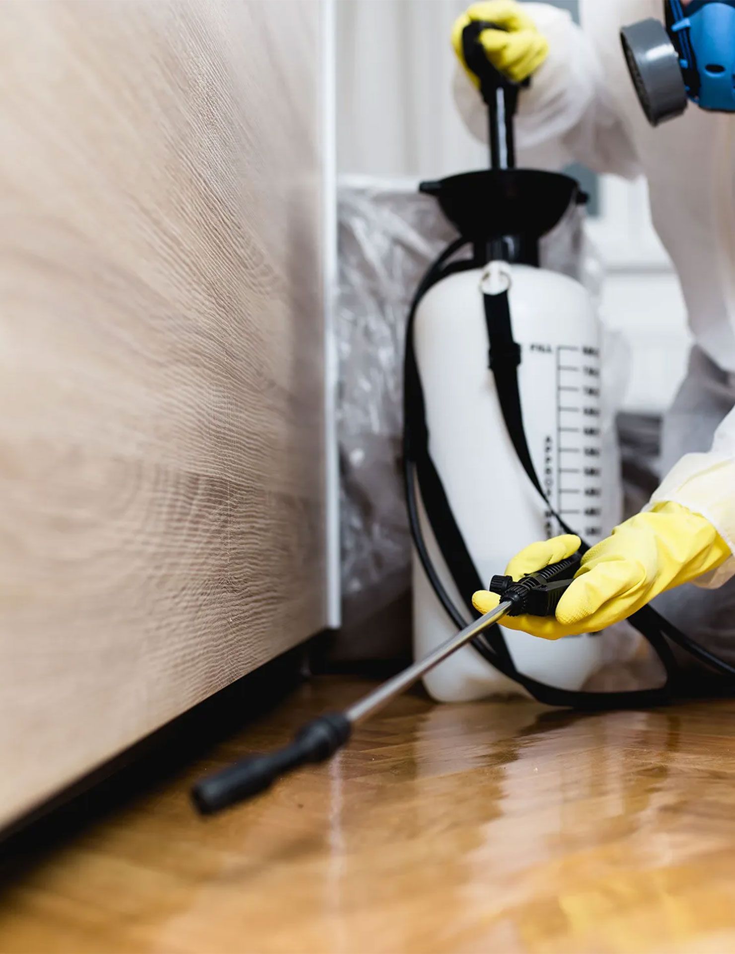 Technician in protective suit sprays pest control near a bed with a sprayer and gloves