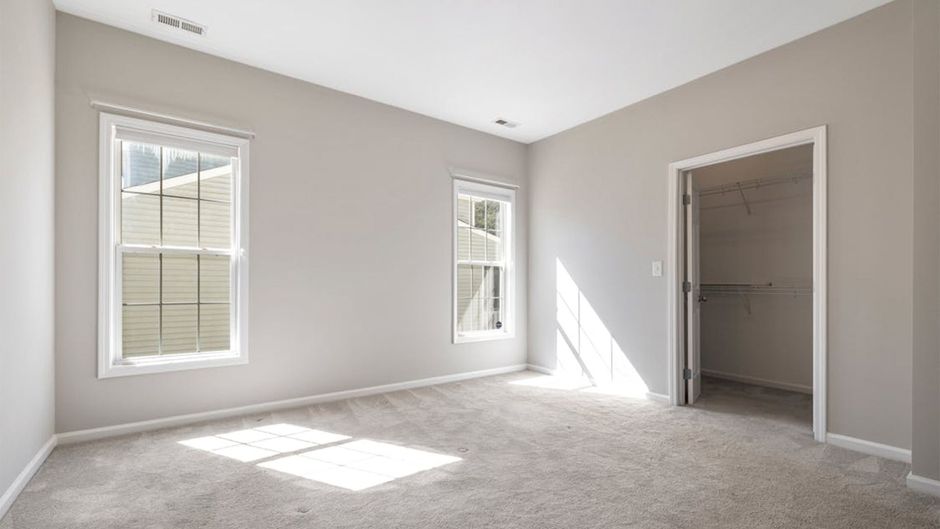 Empty beige bedroom with cleaned carpet, two windows, and an open closet door letting in sunlight