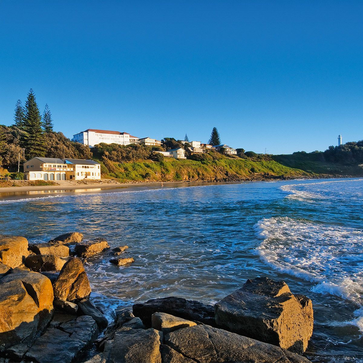 Rocky shoreline with blue waves, coastal houses, and a clear sky at sunset in Yamba