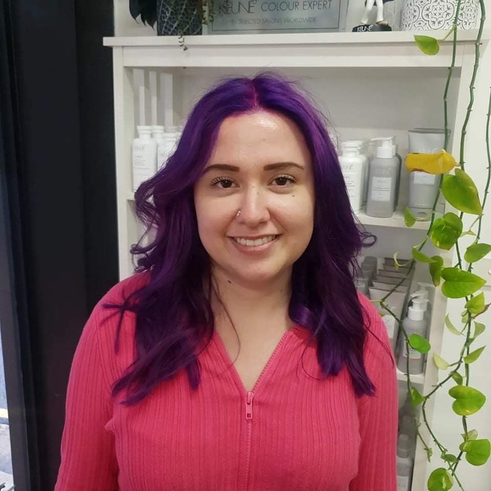 A Woman With Purple Hair is Smiling in Front of a Keune Colour Expert Shelf — Innovative Hair Salon in Gosford, NSW