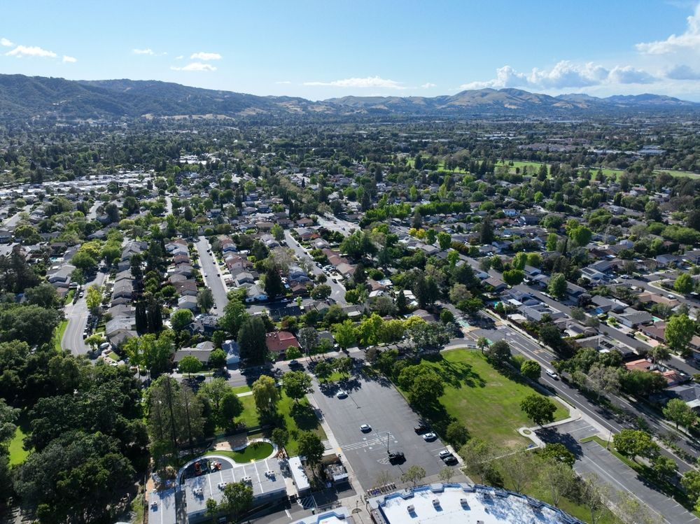Aerial view of suburban neighborhood with green trees, homes, and distant hills under a blue sky.