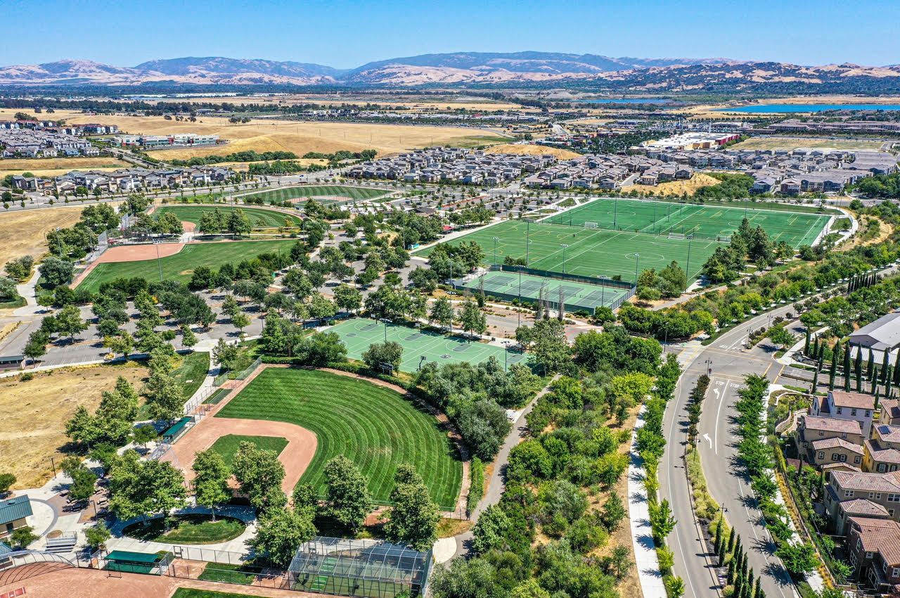 Aerial view of a park with baseball fields, tennis courts, and soccer fields, with residential area in the background.