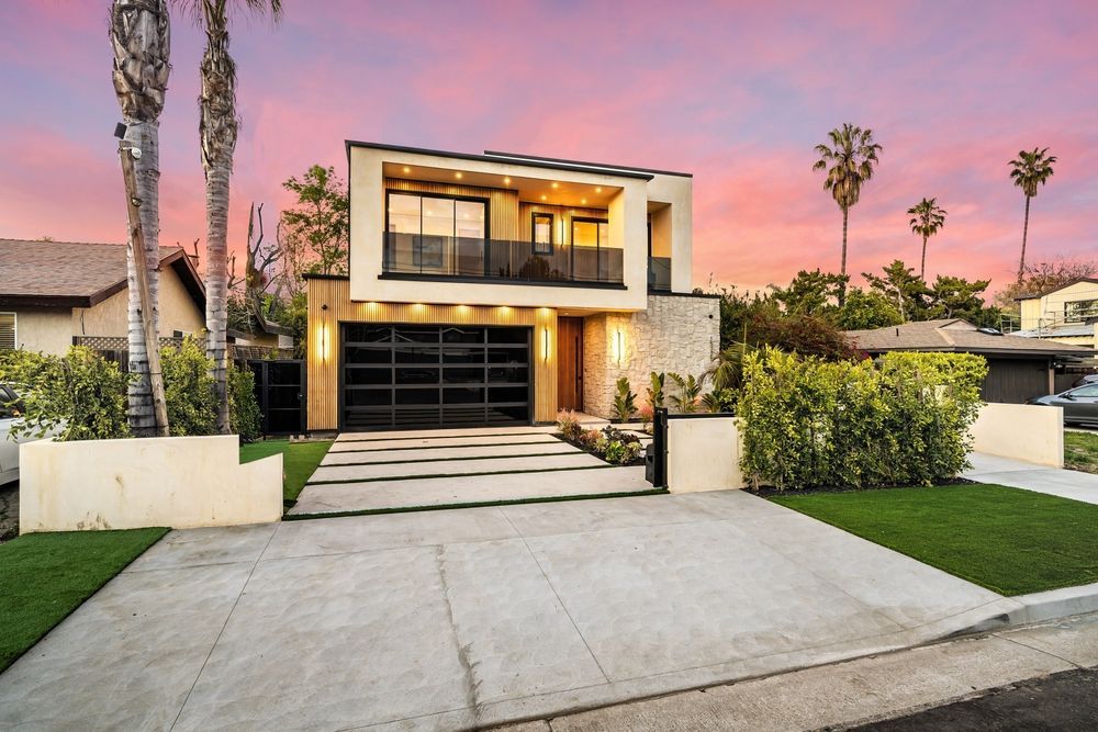 Modern two-story house with black garage door, balcony, and stone accents at sunset with palm trees.