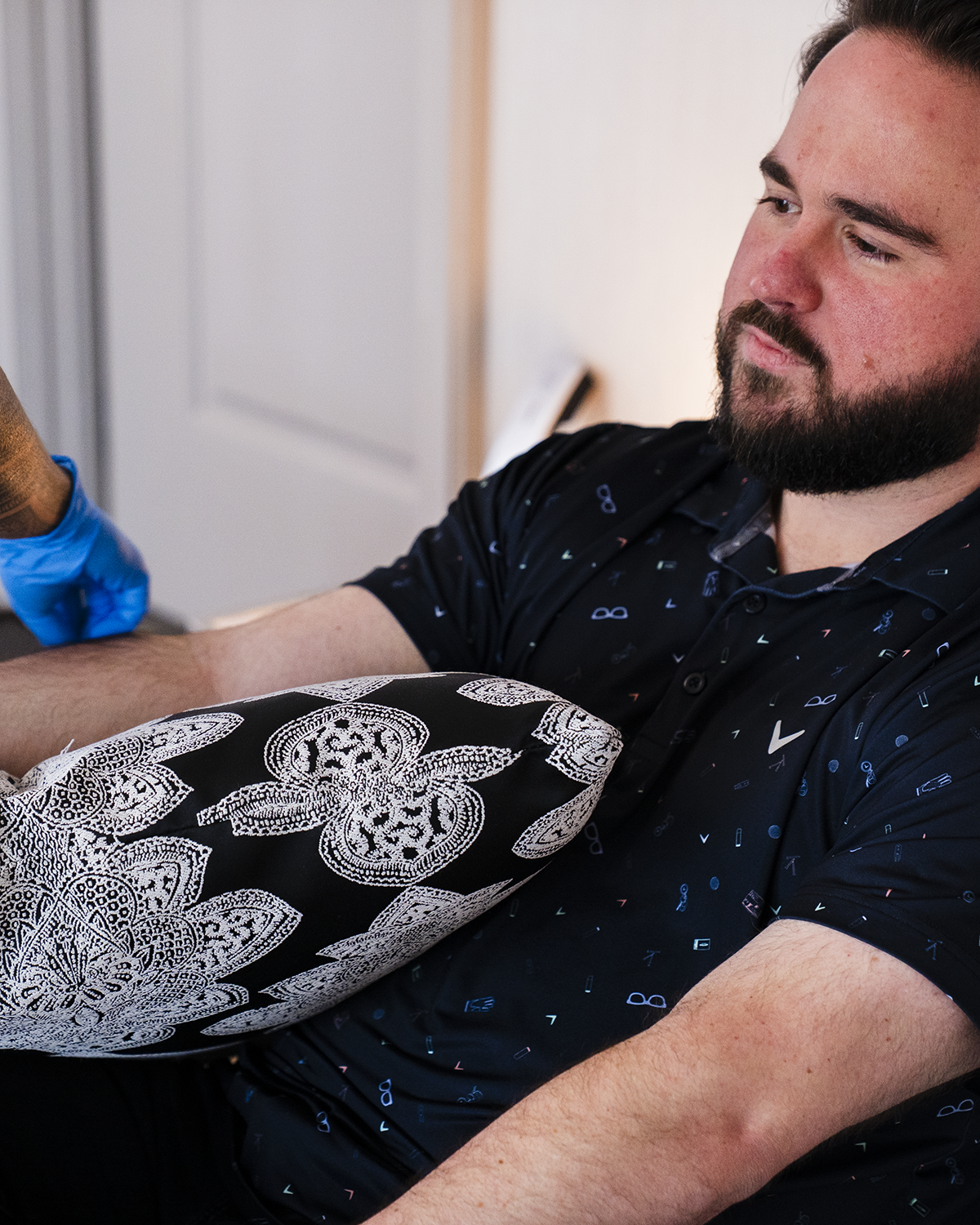 Man in a black shirt with white pattern, looking at his arm while wearing a blue glove and showing a tattooed leg