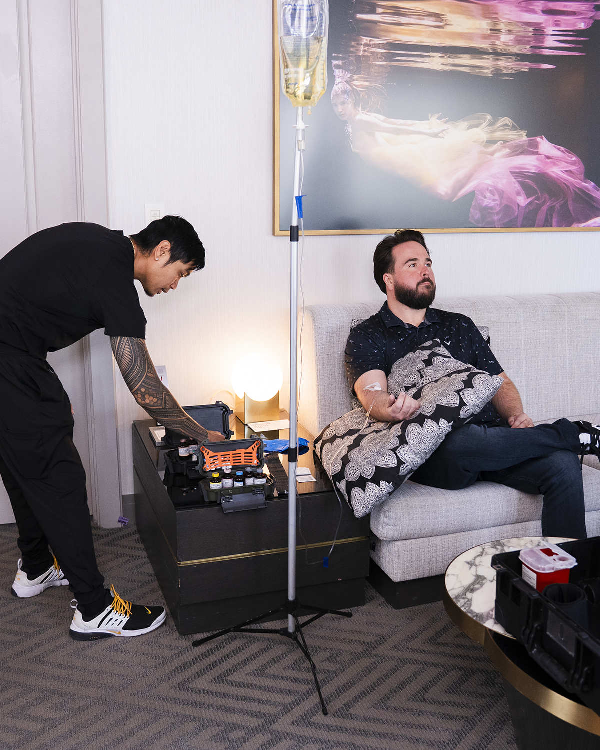 A man adjusts equipment beside a couch while another man relaxes under a blanket in a living room.