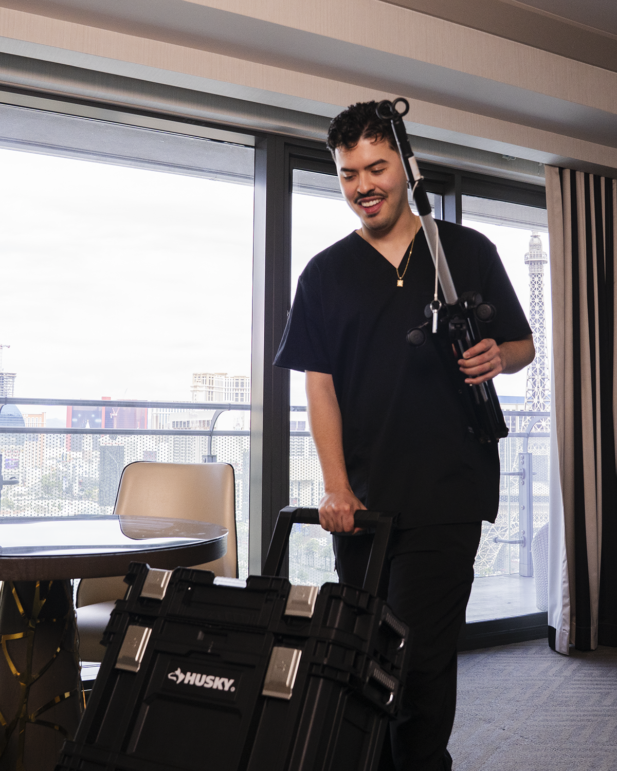 A smiling person in black clothing standing in an office, holding a headset and a small camera beside a table and chair.