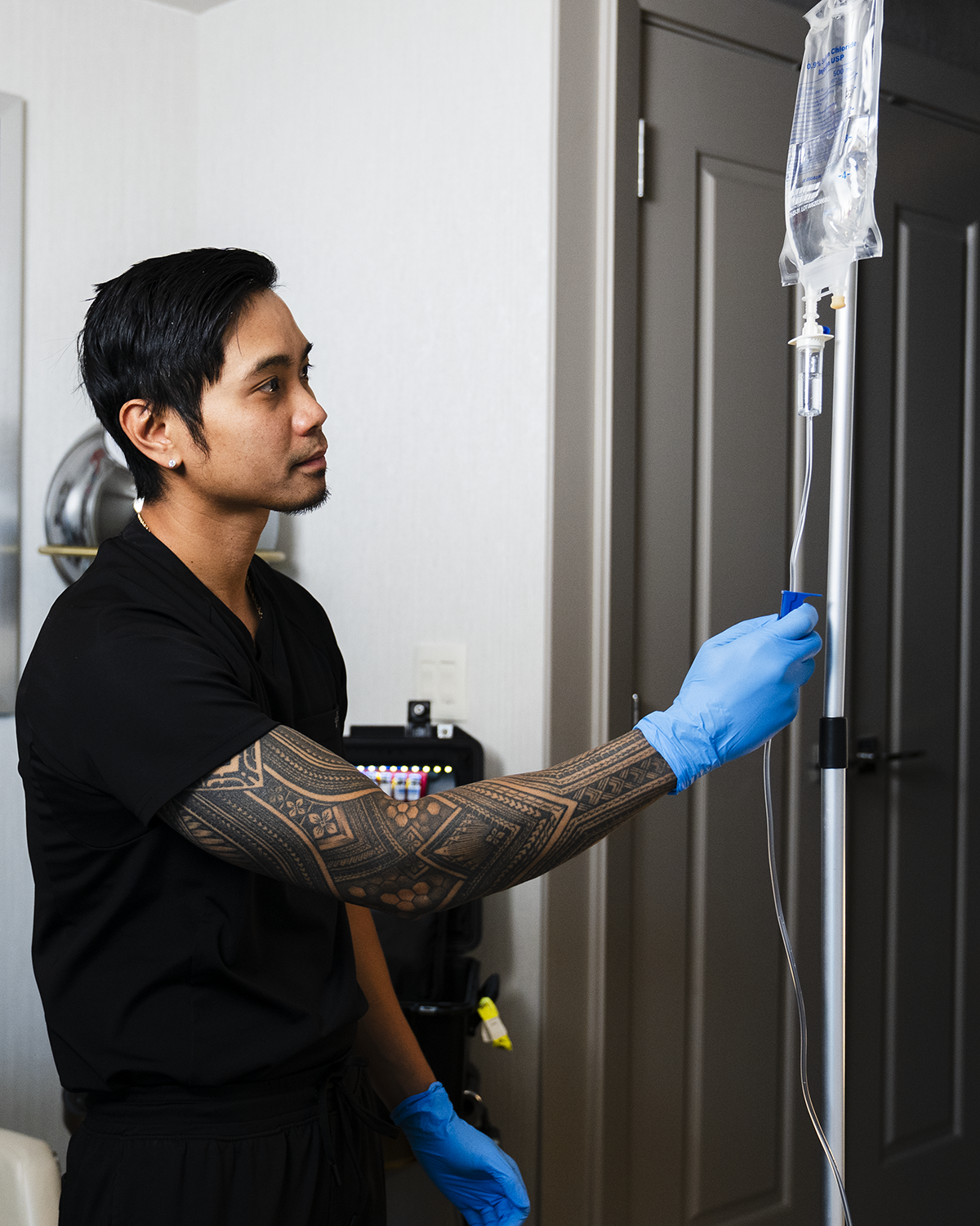 Person in blue gloves hangs an IV bag on a pole beside a dark door.