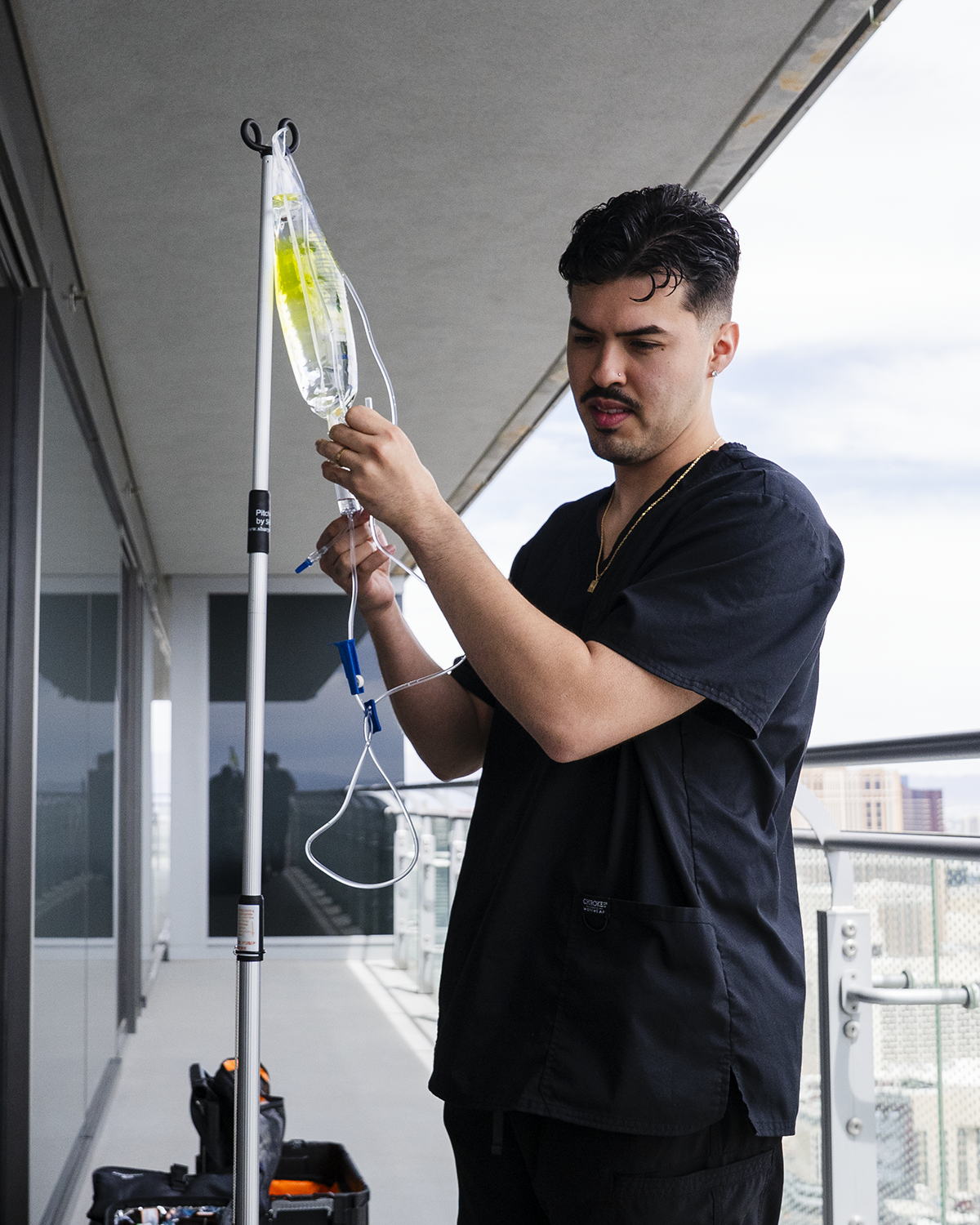 Man in black shirt adjusting an IV drip stand on a balcony with city buildings in the background