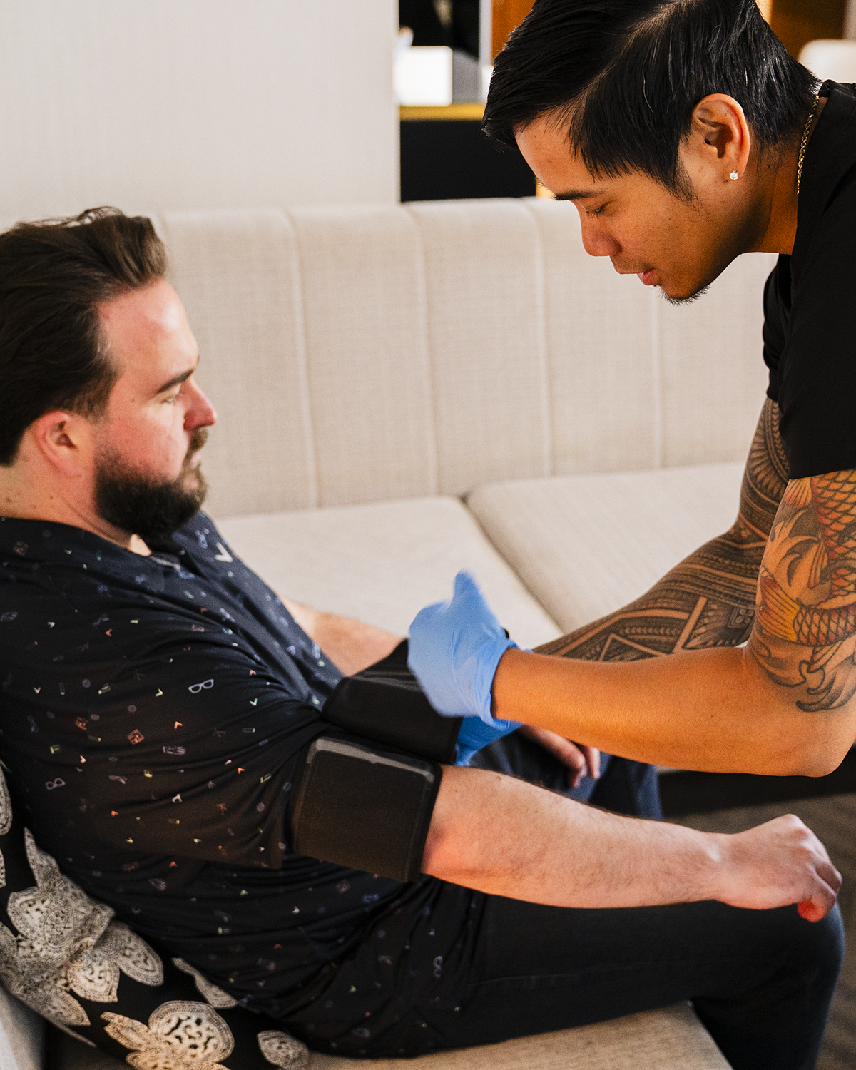 Caregiver in blue gloves fitting a black blood pressure cuff on a seated man’s arm indoors