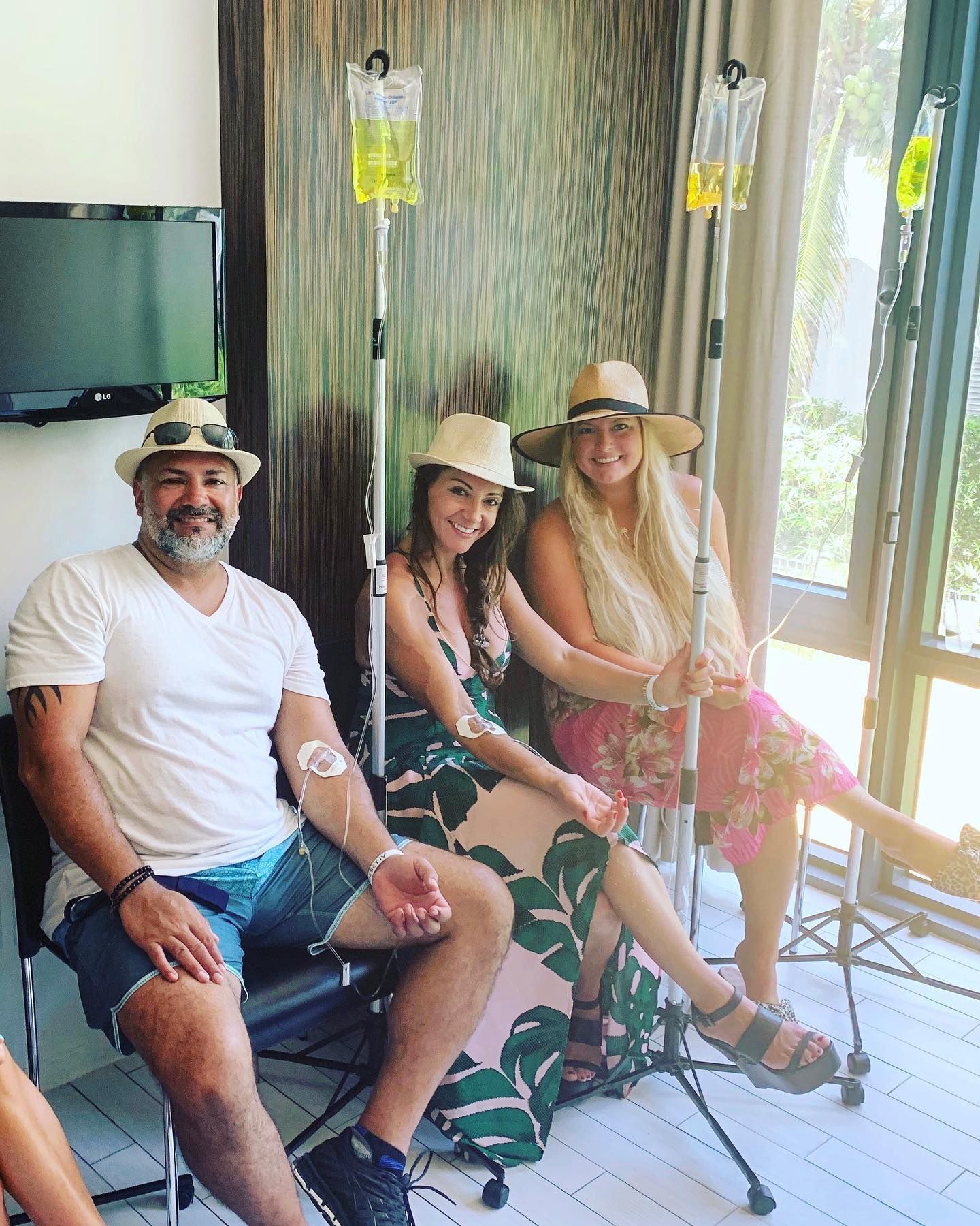 Three people in hats sitting on a porch with a TV and sliding glass door behind them.