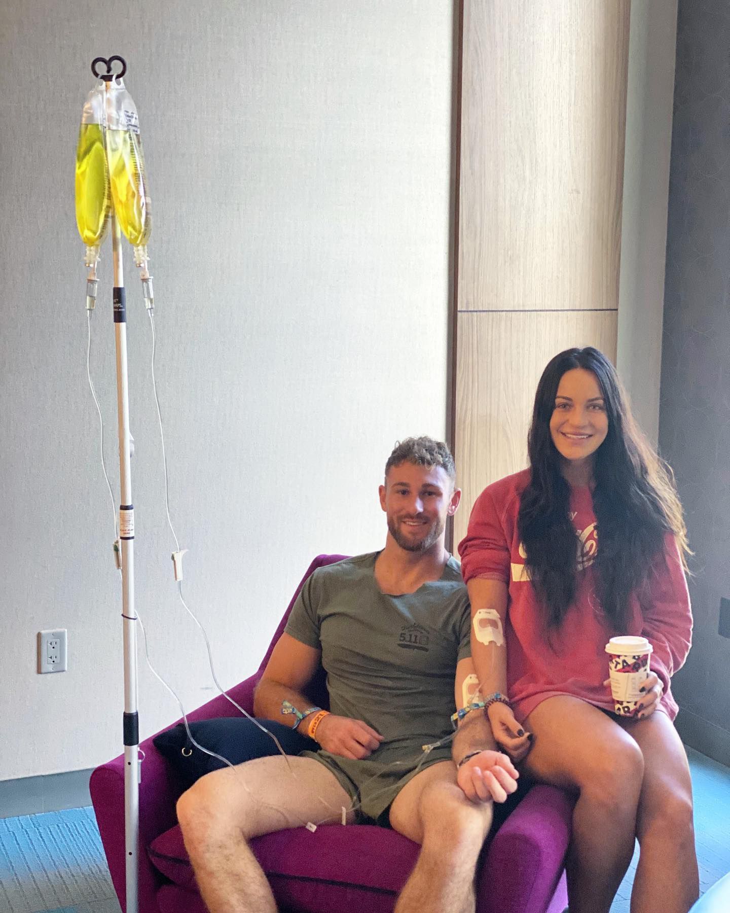 Two seated people smiling beside an IV stand in a hospital room