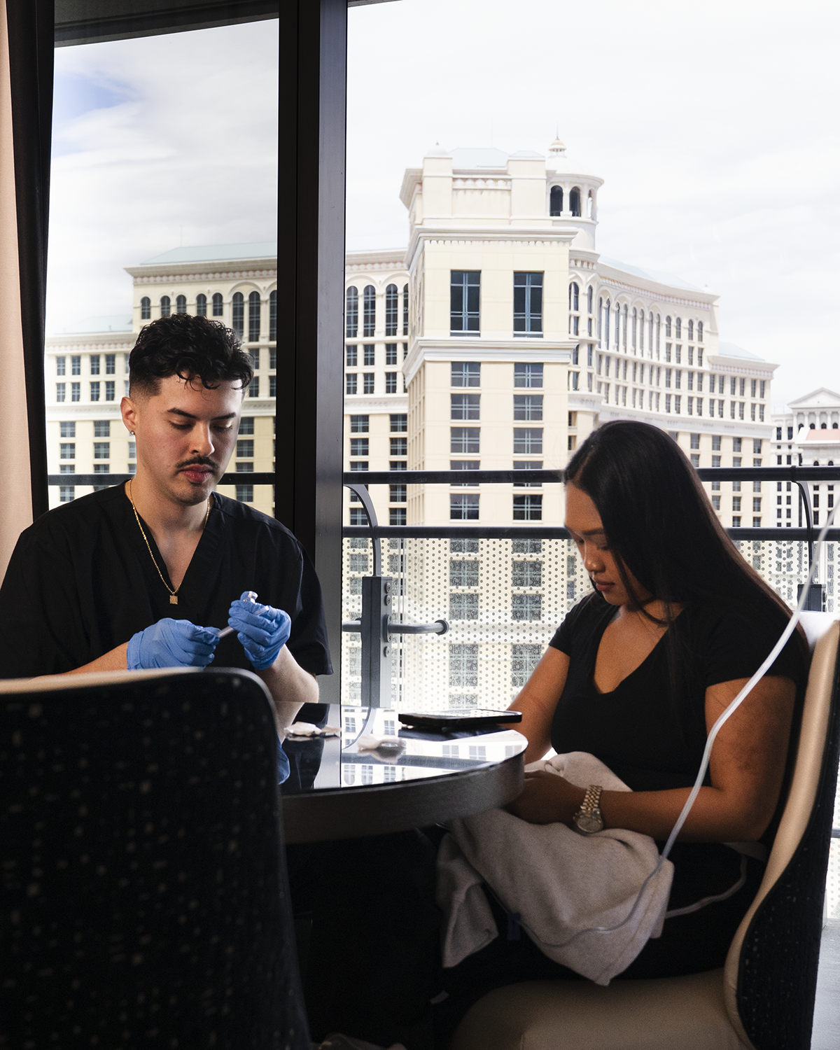 Two people seated at a table by a window, talking while one wears blue gloves in a bright city-view room.