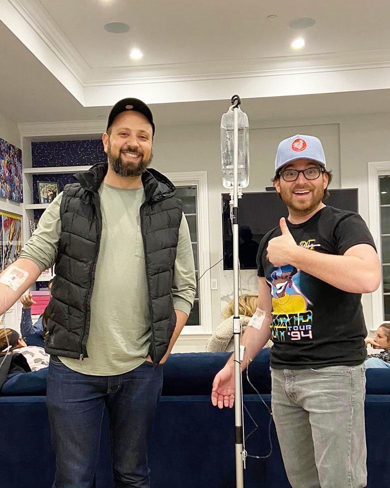 Two men smiling in a living room, one beside a IV pole and the other giving a thumbs-up.