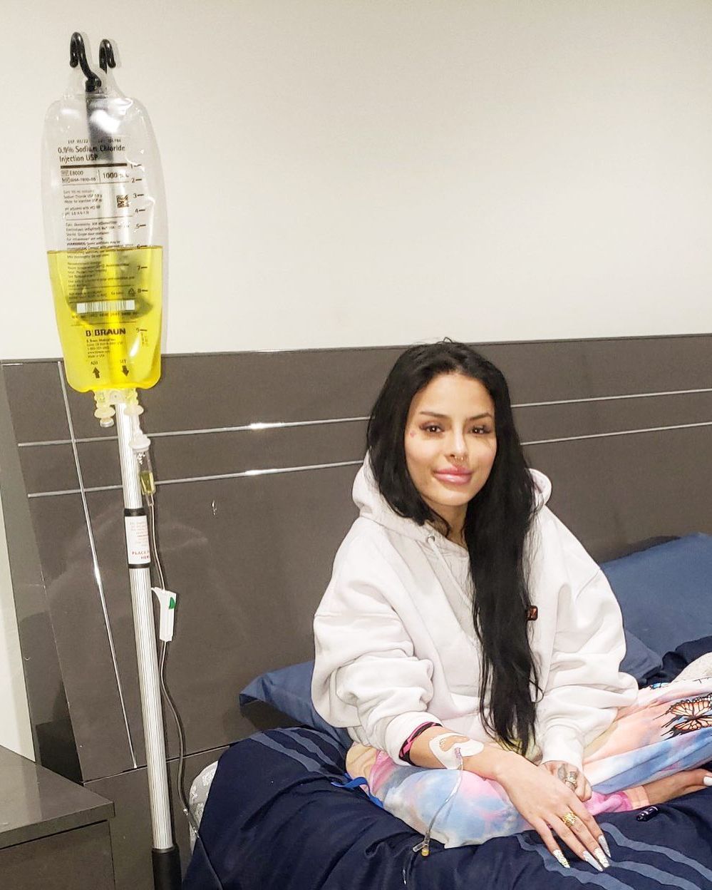 Patient seated in bed beside an IV drip, wearing a white sweatshirt and smiling at the camera.