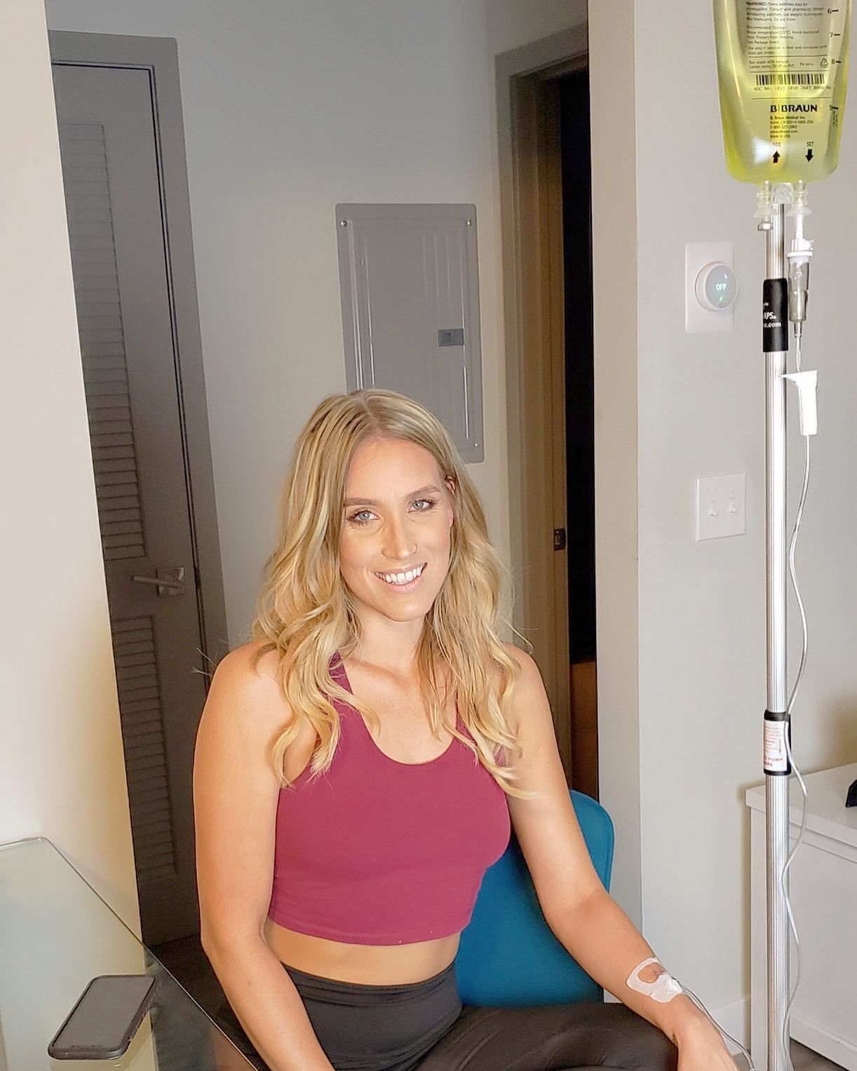 Woman smiling beside an IV stand in a home setting, wearing a pink crop top and black pants.