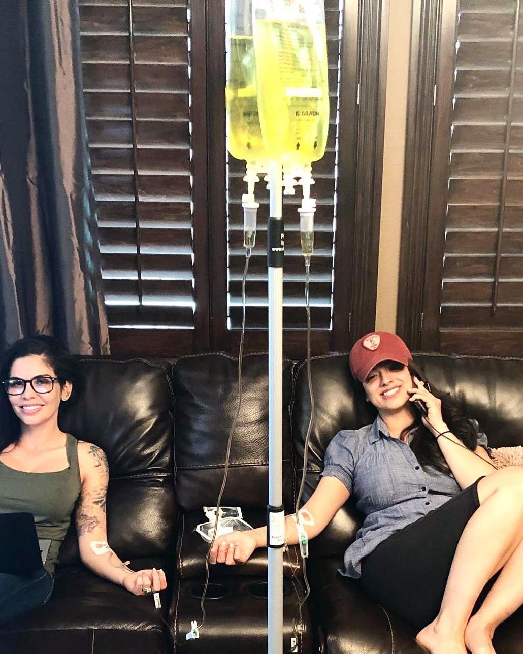 Two people sit on a black couch beside an IV stand in a dim room with wooden shutters.