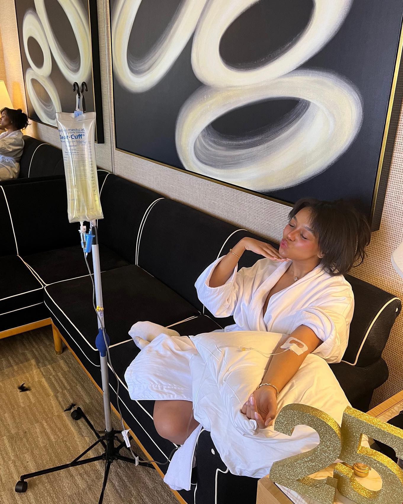 Person in white clothing sits on a couch beside an IV stand in a modern lounge.