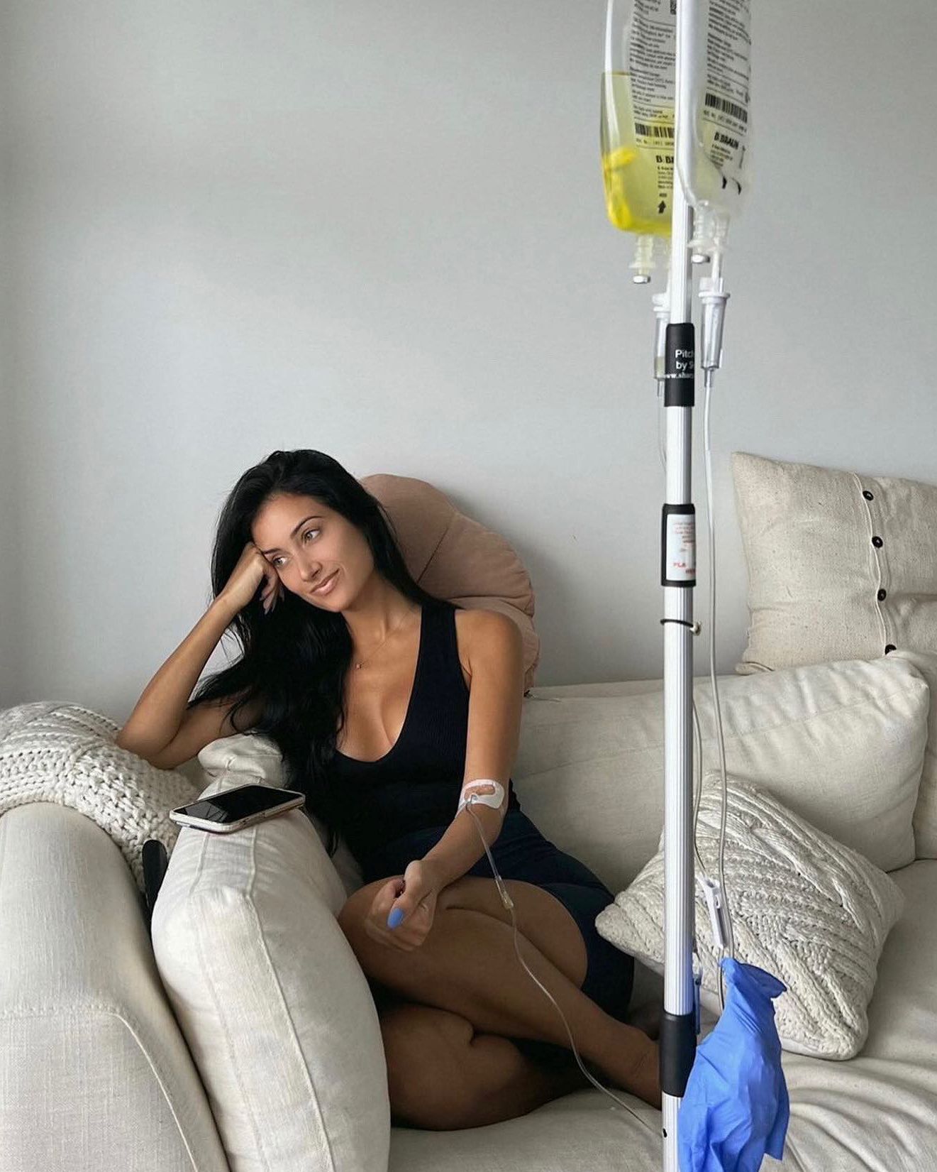 Woman sitting on a couch beside an IV stand, resting with a blanket in a bright living room.