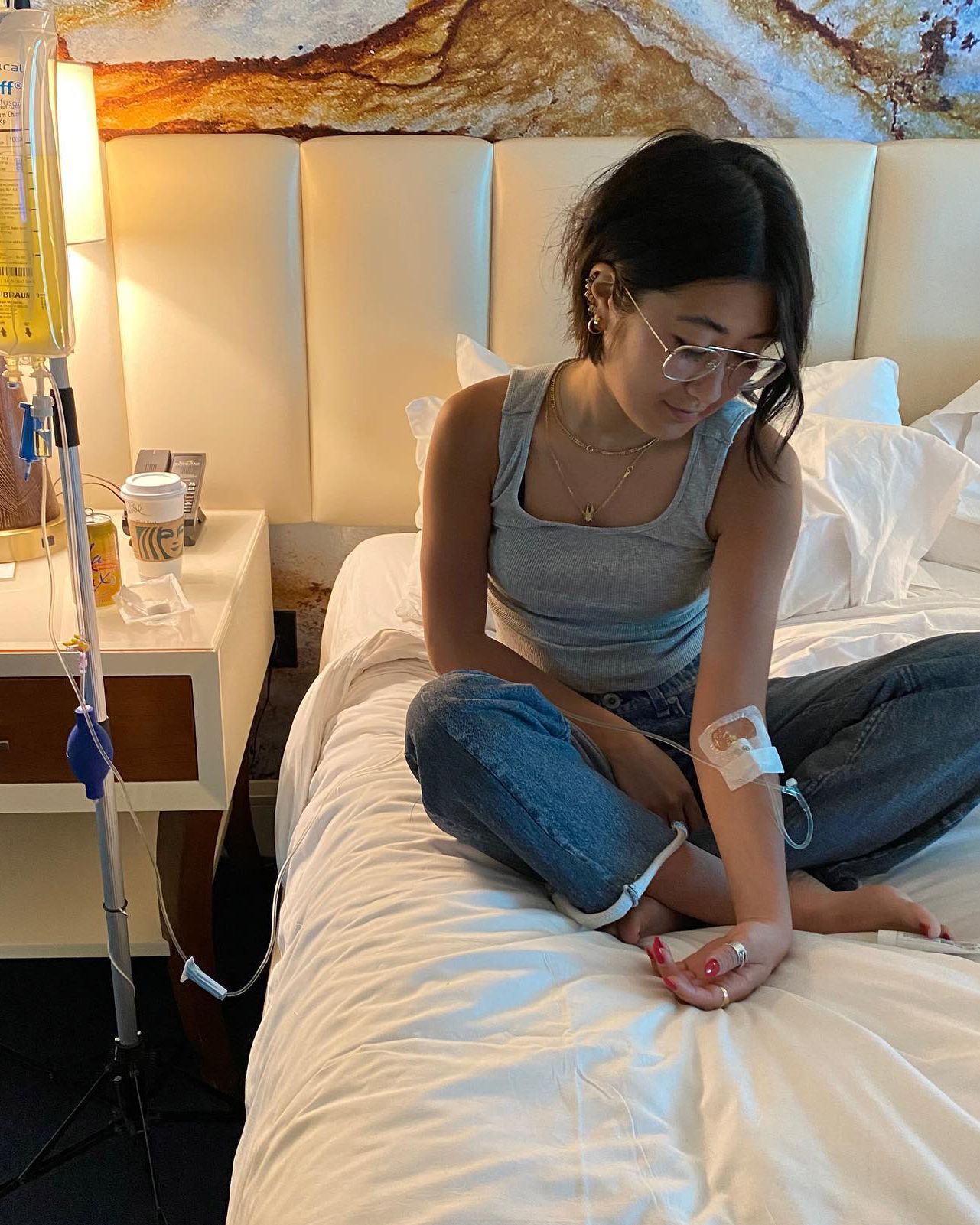 Person sitting cross-legged on a bed, looking down at a phone in a softly lit hotel room.