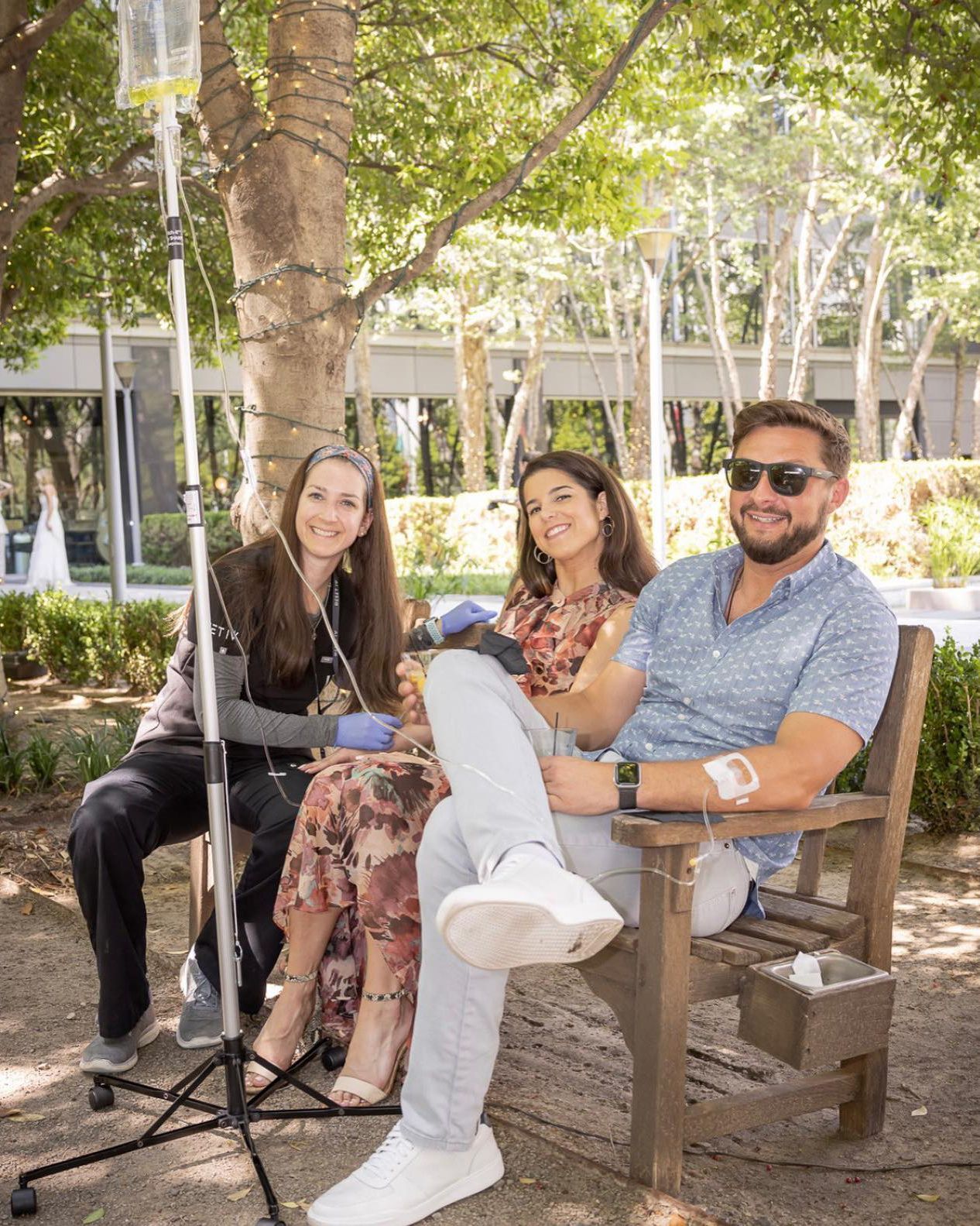 Three people sitting on a bench under a tree, smiling in a sunny outdoor courtyard.