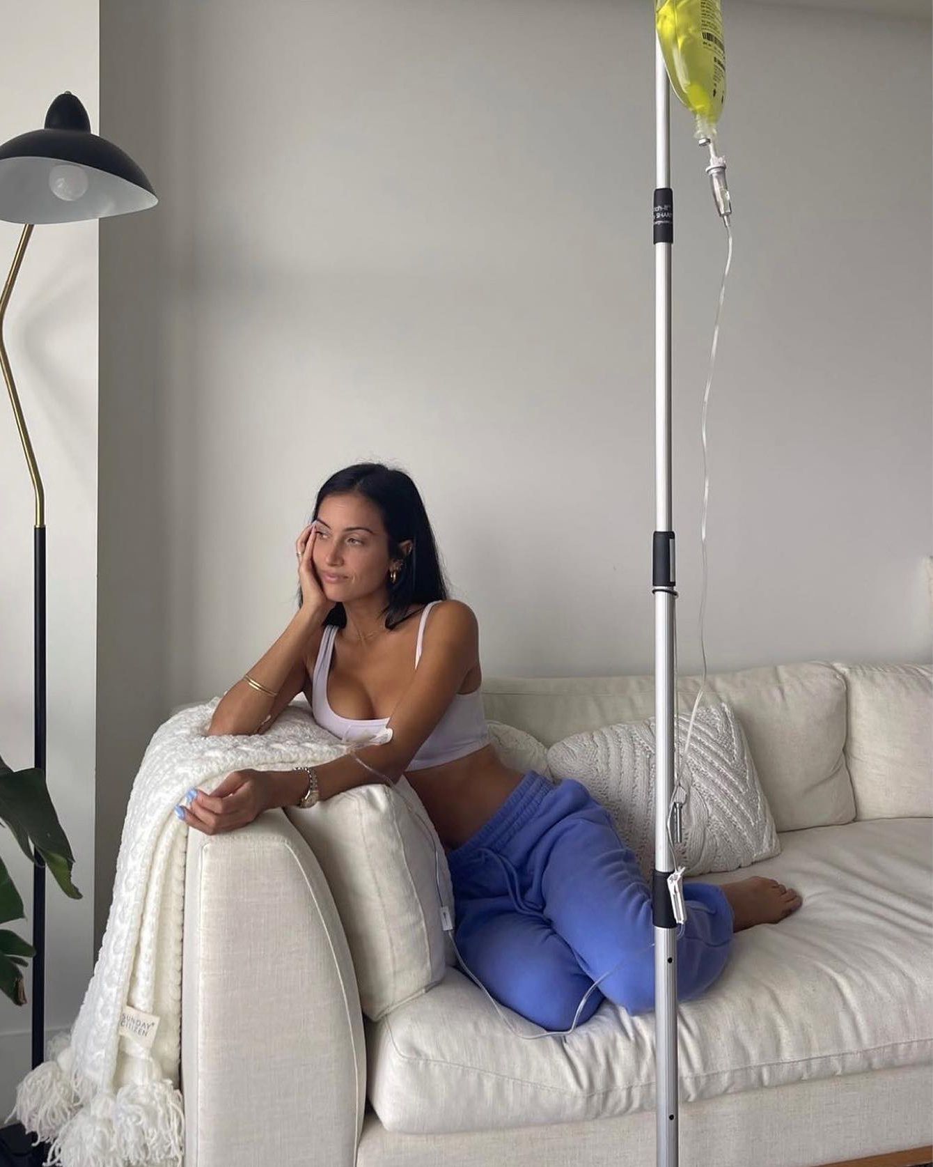 Woman reclining on a white sofa in a bright living room beside a floor lamp and houseplant.