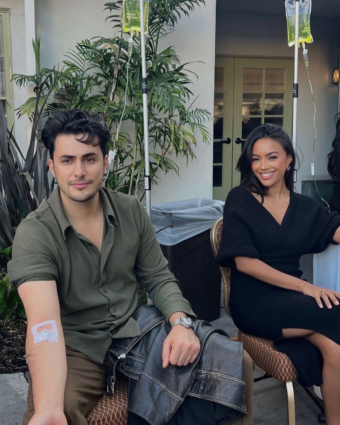 Two people seated outdoors on a patio with plants and hanging lights, smiling at the camera.