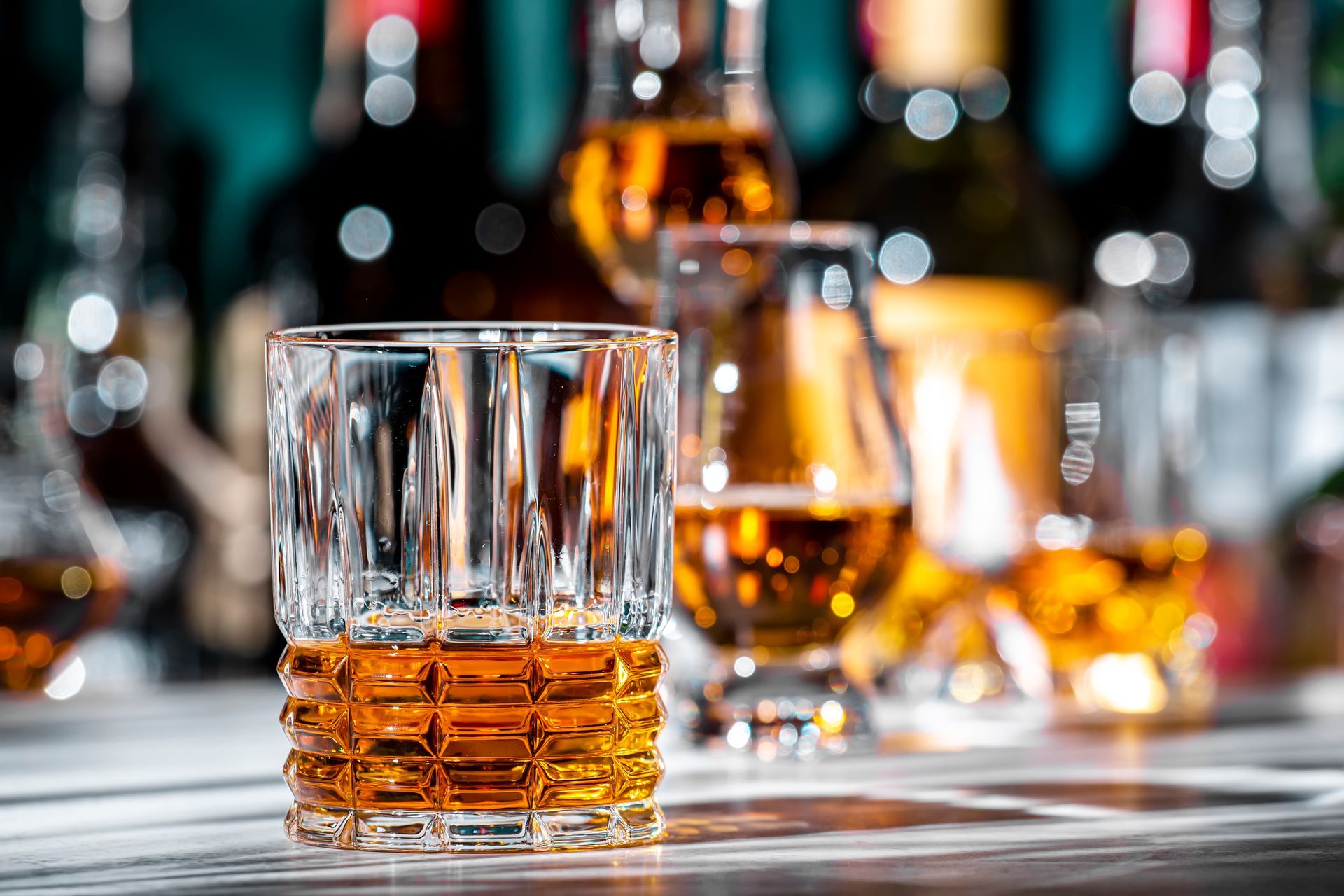 Two whiskey glasses with amber liquor on a bar counter, with blurred bottles and lights in the background