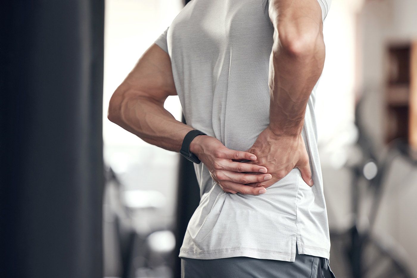 Person holding lower back in gym, wearing a white shirt and gray shorts