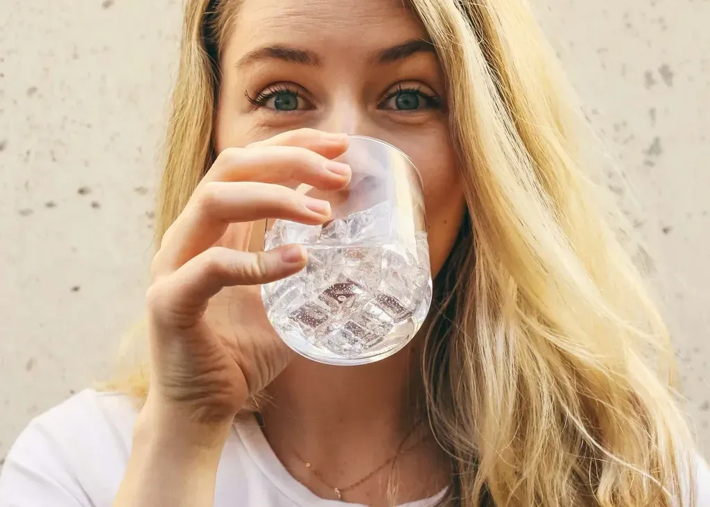 Woman with blonde hair drinking water from a glass, smiling at the camera outdoors.