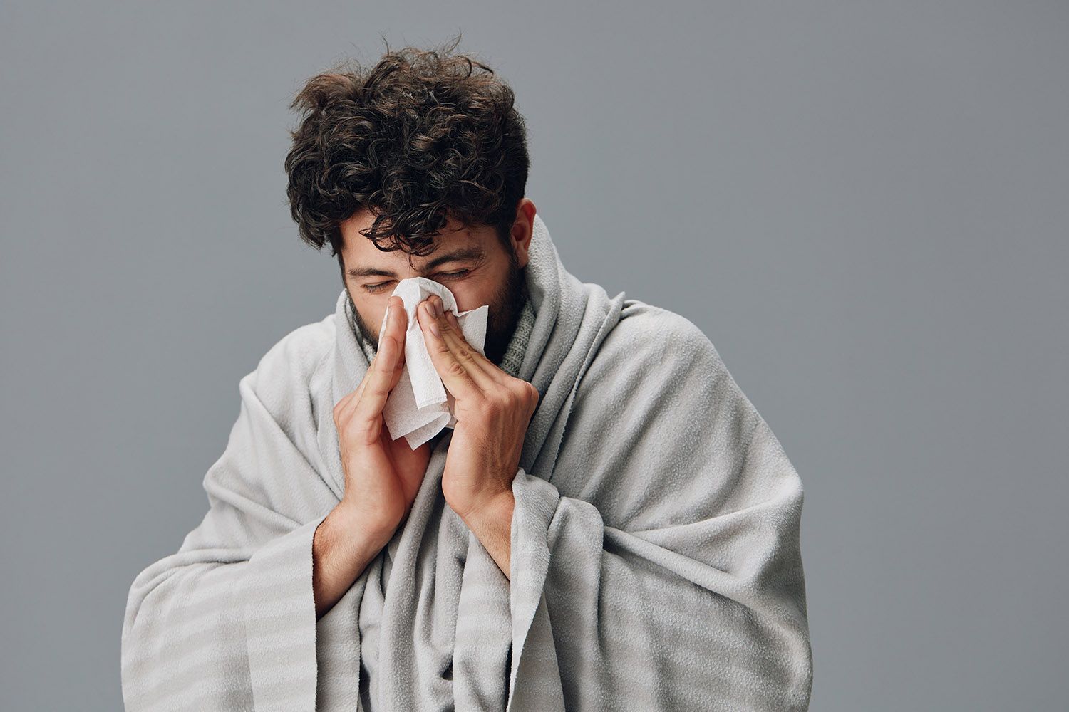 Man in a gray robe holding a tissue to his nose against a plain gray background