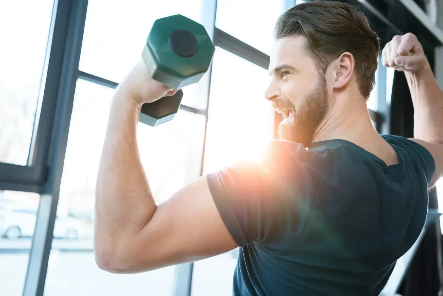 Man lifting dumbbells in a bright gym, flexing biceps with a sunlight flare.