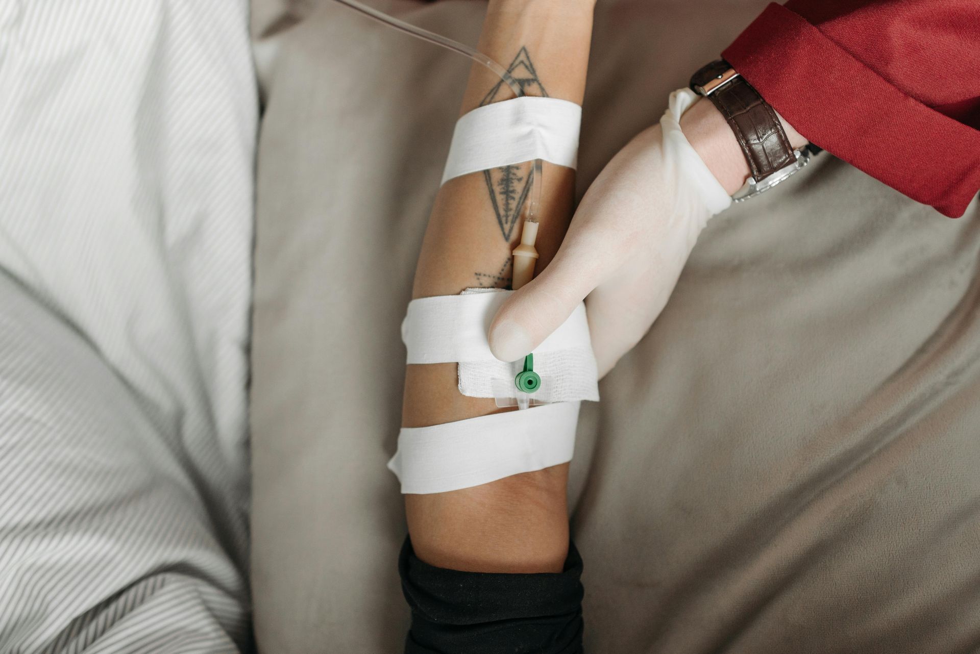 Gloved hands inserting an IV line into a tattooed arm, secured with tape