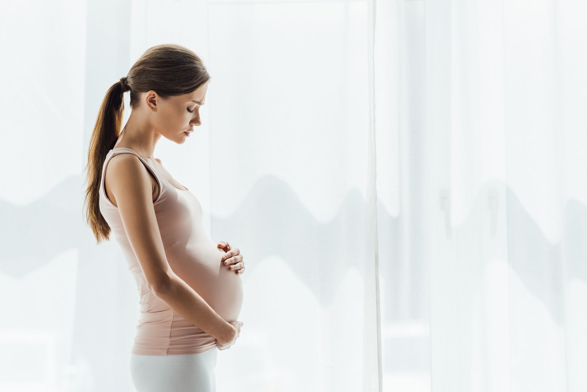 Pregnant woman in profile, standing by a bright window with hands on her belly