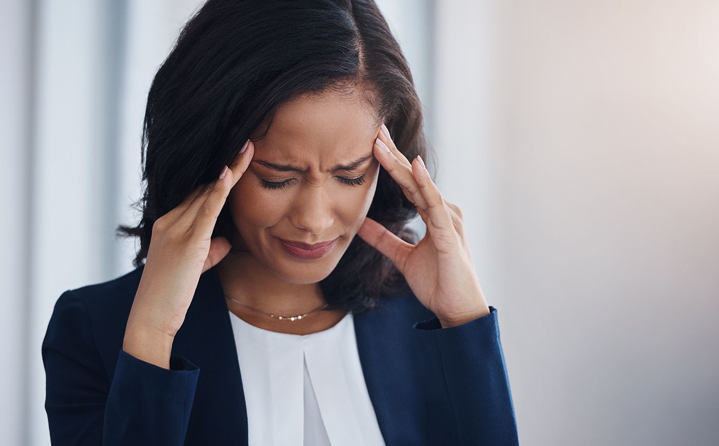 Woman in a dark blazer holding her temples with a pained expression, appearing to have a headache.
