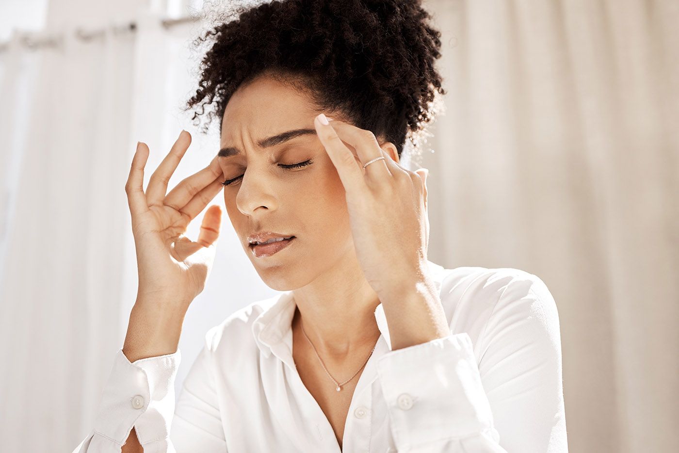 Woman in a white shirt massaging her temples with eyes closed, looking stressed