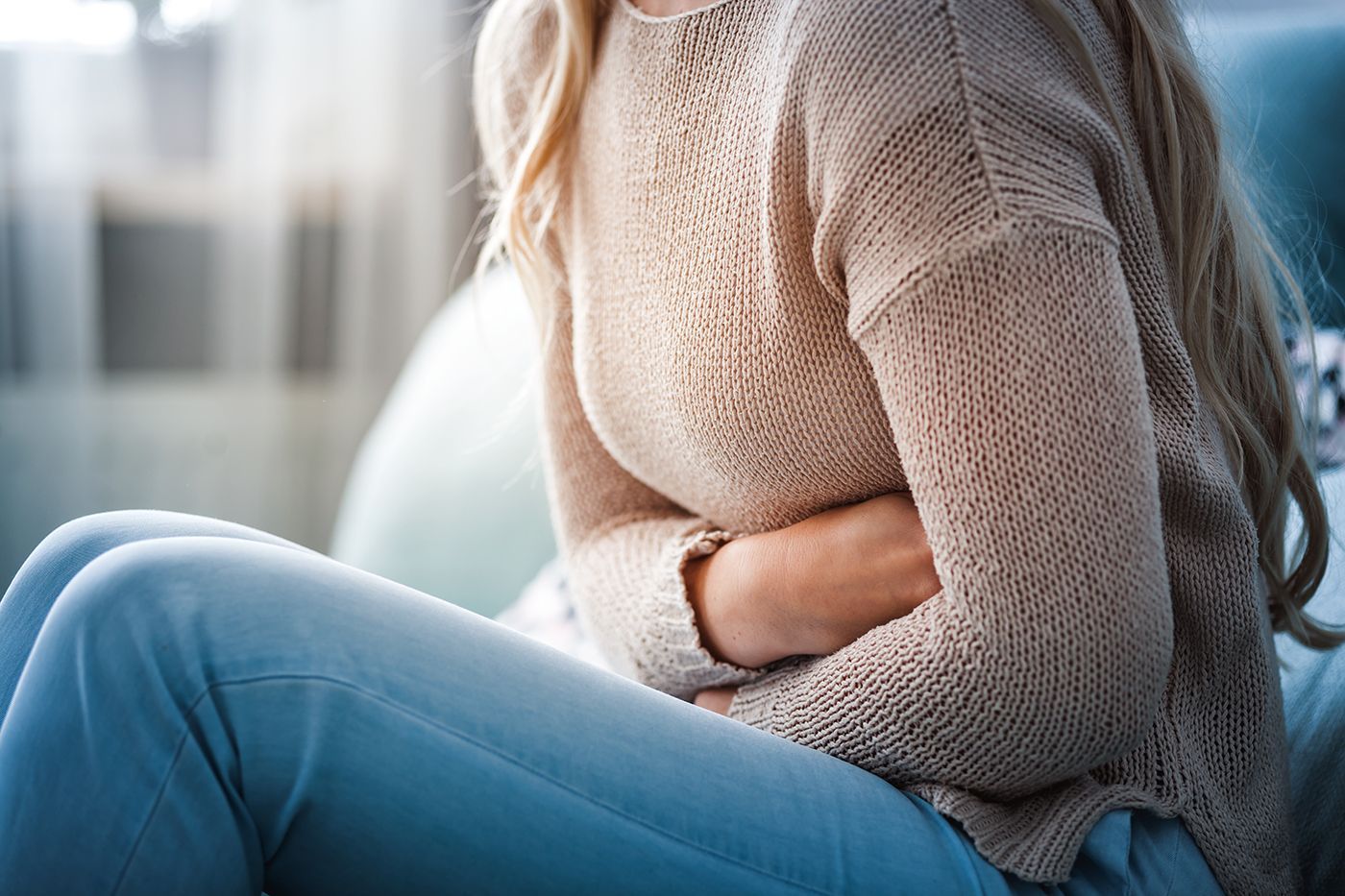 Person sitting on a couch, clutching their stomach in discomfort, wearing a beige sweater and blue jeans.