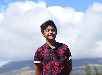 A young man in a floral shirt is standing in front of a cloudy sky.